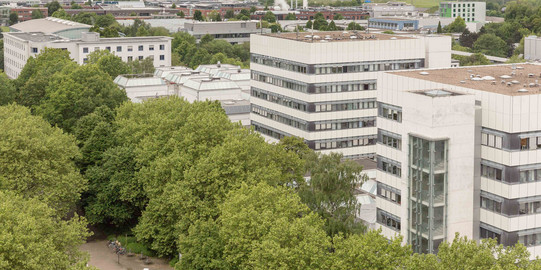 BCI: Rechts im Bild befinden sich zwei hohe graue Gebäude mit vielen Fenstern der Fakultät Bio- und Chemieingenieurwesen. Davor und weiter links im Bild Bäume.