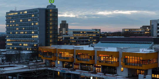 Foto von Mathetower mit grünem TU Logo auf dem Dach und daneben gelegene Mensa beleuchtet im Winter